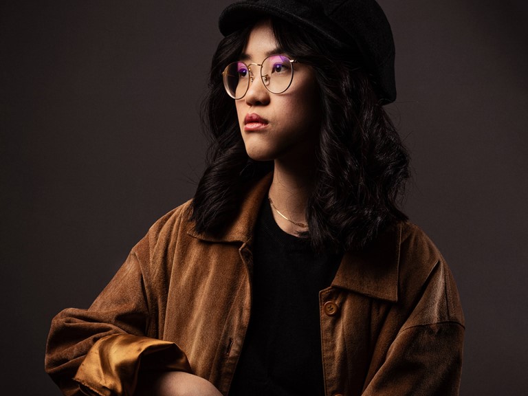 An Asian-looking woman with twisted black hair, large round glasses and a black hat poses in the style of the old masters against a dark background. She is wearing a cognac-coloured oversize jacket.