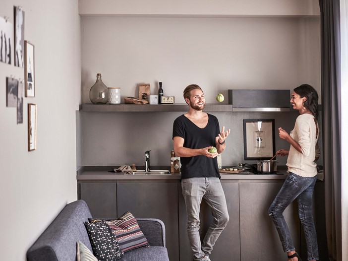 On the left a couch with cushions, in the middle a dark grey kitchenette with accessories, on the right in front a smiling man with a beard in a black T-shirt juggling fruit, on the left a dark-haired woman in a white blouse looking at him with a smile while stirring a pot. 