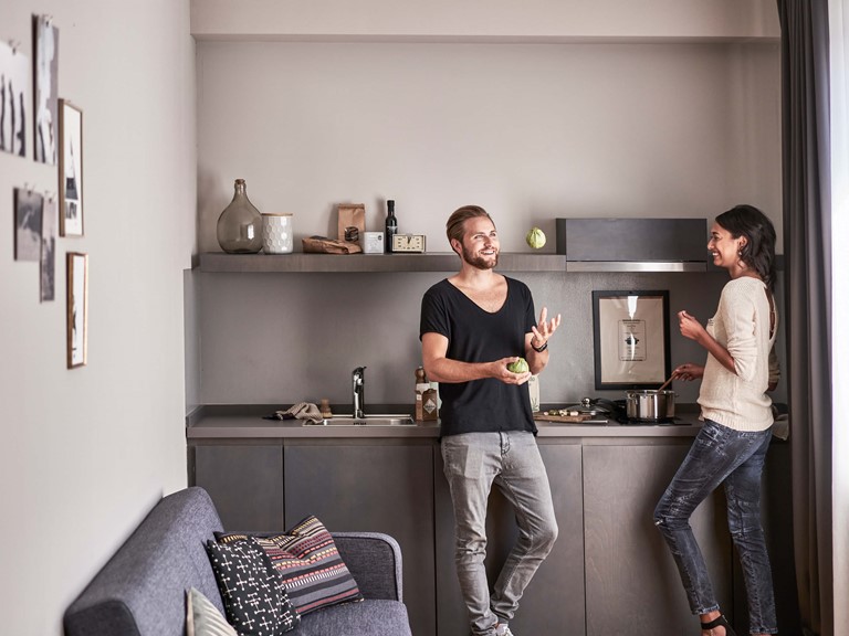 On the left a couch with cushions, in the middle a dark grey kitchenette with accessories, on the right in front a smiling man with a beard in a black T-shirt juggling fruit, on the left a dark-haired woman in a white blouse looking at him with a smile while stirring a pot. 