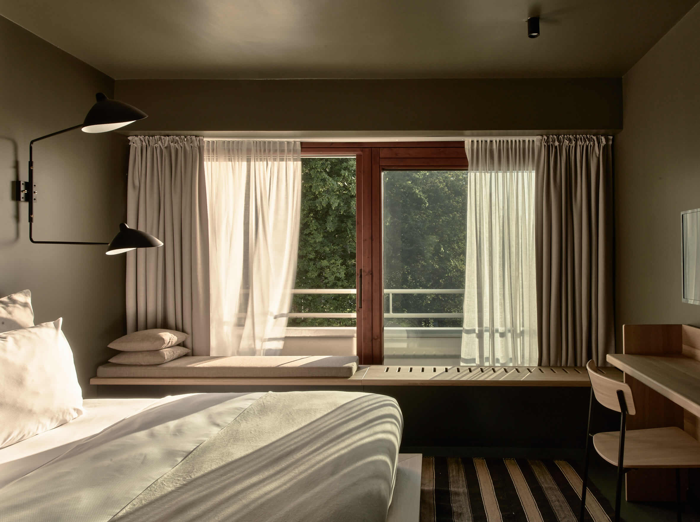 Links ein hell bezogenes Doppelbett, daneben eine schwarze Wandleuchte, rechts ein Schreibtisch aus Holz mit Stuhl. Frontal eine Fensterfront mit Schiebetür zu einem Balkon, links auf der breiten Fensterbank eine Sitzecke.