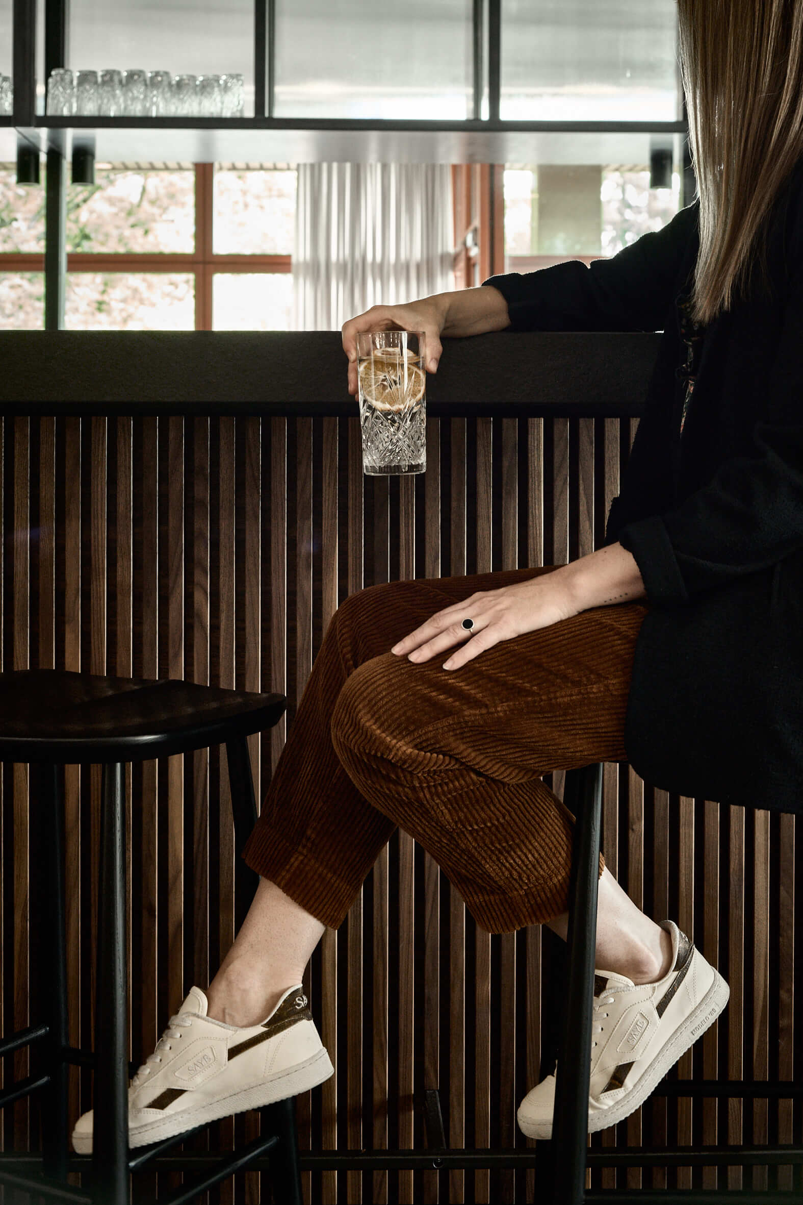 Woman on a stool at a bar with a wooden counter, she is wearing a black blazer, brown corduroy trousers and trainers, leaning casually on the bar with a drink in her hand. 