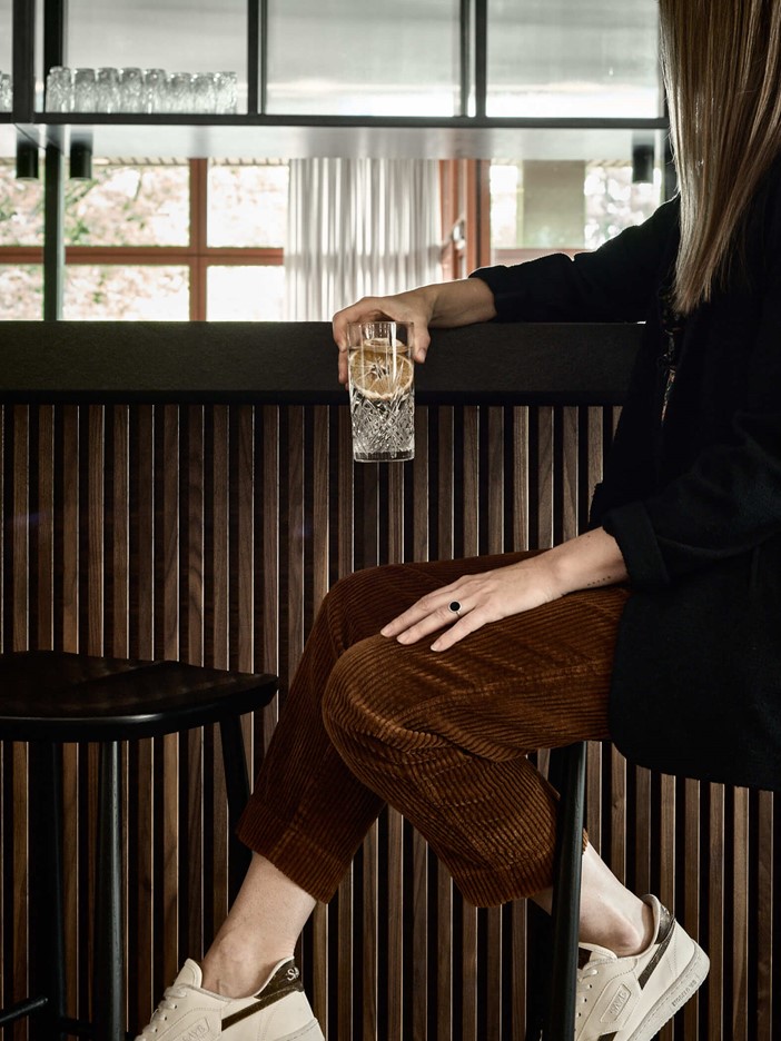 Frau auf einem Hocker an einer Bar mit hölzernem Tresen, sie trägt einen schwarzen Blazer, eine braune Breitcordhose und Turnschuhe, stützt sich lässig auf der Bar ab mit einem Drink in der Hand. 