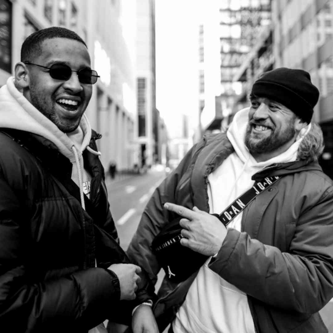 Bruno Banarby and Halid Rizvanovic - two men - are laughing. They are standing in a street. One of the men is wearing a black jacket and sunglasses. He has short hair. The man on the right is wearing a bomber jacket, a fanny pack and a cap.