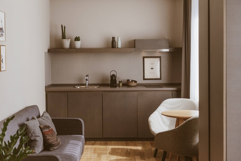 Zimmer mit hellem Holzboden und einer dunkelgrauen Küchenzeile, linker Hand eine graue Couch mit Kissen, rechts ein runder Holztisch mit Sesseln an einem großen Fenster.