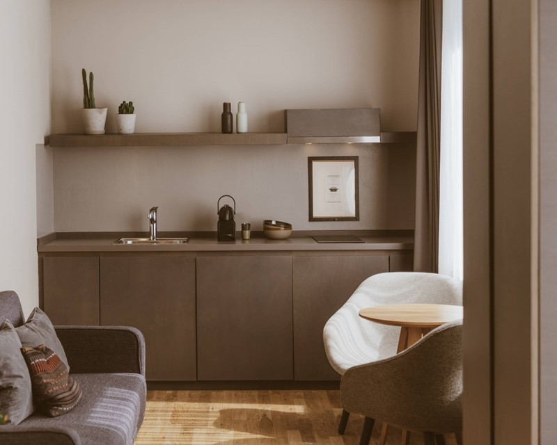 Zimmer mit hellem Holzboden und einer dunkelgrauen Küchenzeile, linker Hand eine graue Couch mit Kissen, rechts ein runder Holztisch mit Sesseln an einem großen Fenster.
