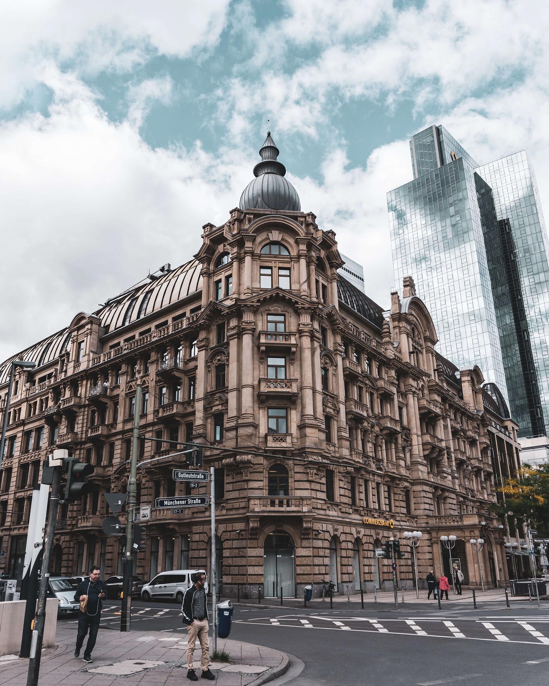 Corner of a street with a man standing and waiting, a jogger approaching, a historic-looking corner building and a modern skyscraper across from it