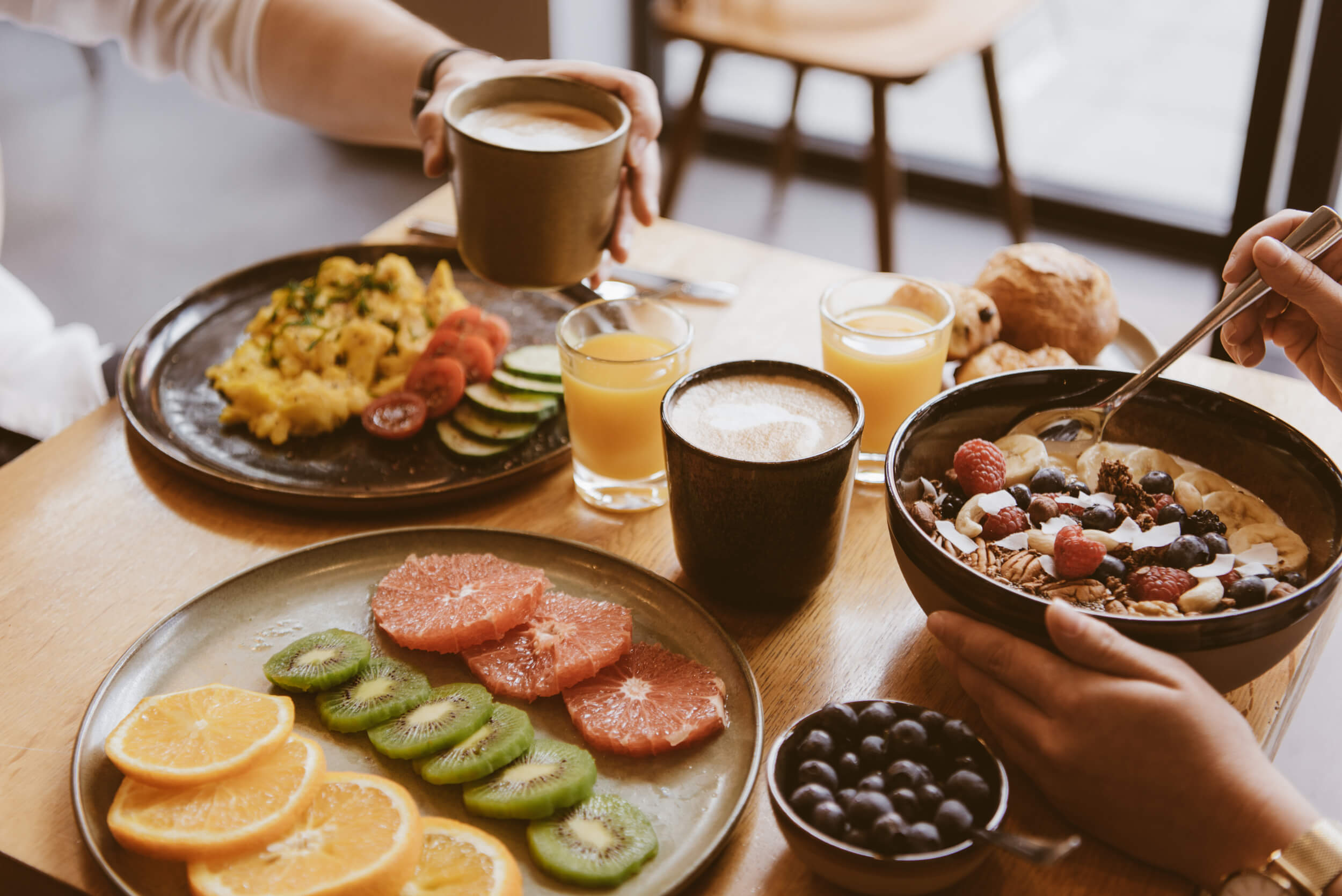 Coffee, orange juice, porridge in a bowl, scrambled eggs with vegetables, pastries and beautifully arranged fruit on a light wooden table at which two people are sitting, with only their hands visible as they have breakfast.  