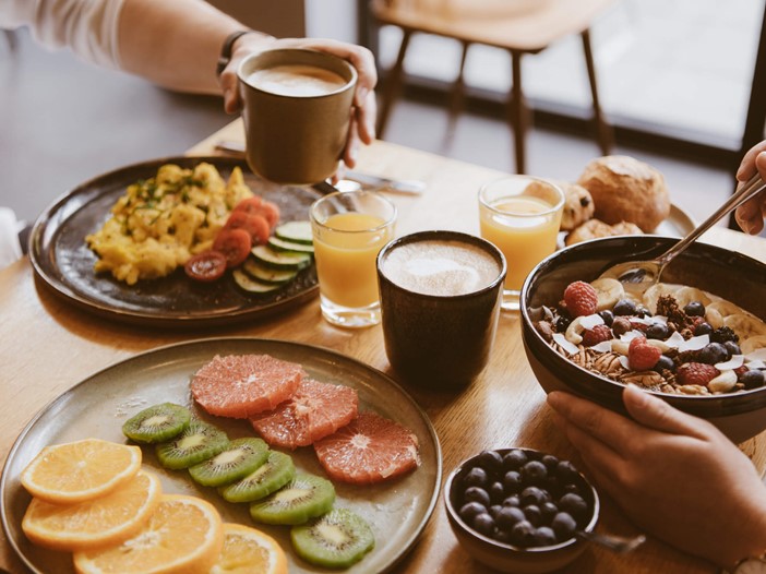Kaffee, Orangensaft, Porridge in einer Schale, Rührei mit Gemüse, Gebäck und schön angerichtetes Obst auf einem hellen Holztisch, an dem zwei Personen sitzen, nur deren Hände beim Frühstücken sind zu sehen.  