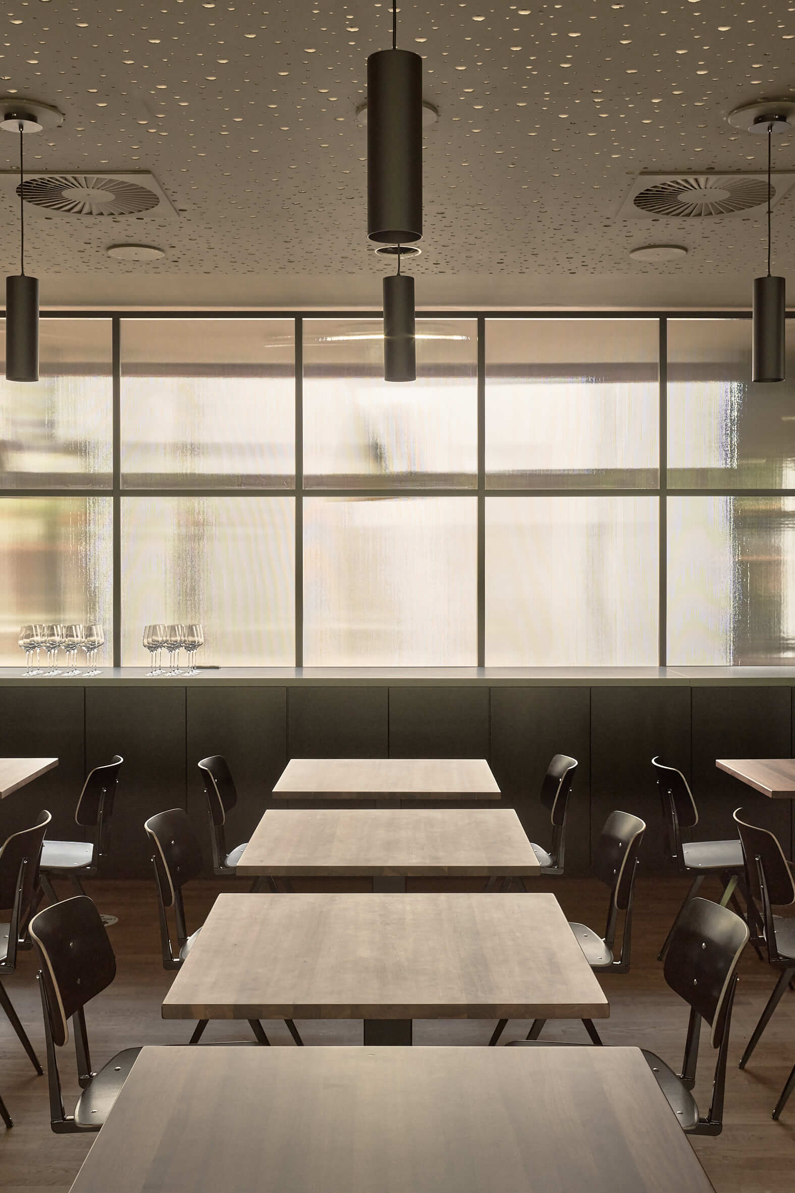 Restaurant mit dunklen Stühlen an kleineren Holztischen. Den Hintergrund bestimmt eine Milchglaswand, die viel Licht herein lässt.