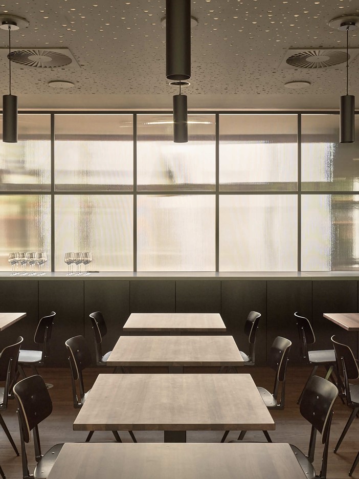 Restaurant with dark chairs at smaller wooden tables. The background is defined by a frosted glass wall that allows a lot of light into the room.