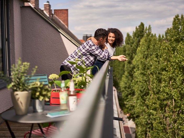 A roof terrace with a table, plants and colourful chairs, A man wearing a checked shirt leans on the railing, a woman with dark hair is facing him and embraces him smiling. 
