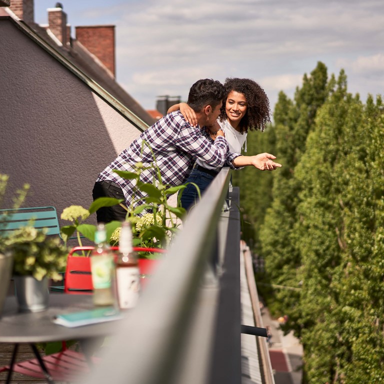 Eine Dachterrasse mit Tisch, Pflanzen und bunten Stühlen, Ein Mann mit kariertem Hemd lehnt sich auf das Geländer, eine Frau mit dunklen Haaren ist ihm zugewandt und umarmt ihn lächelnd. 