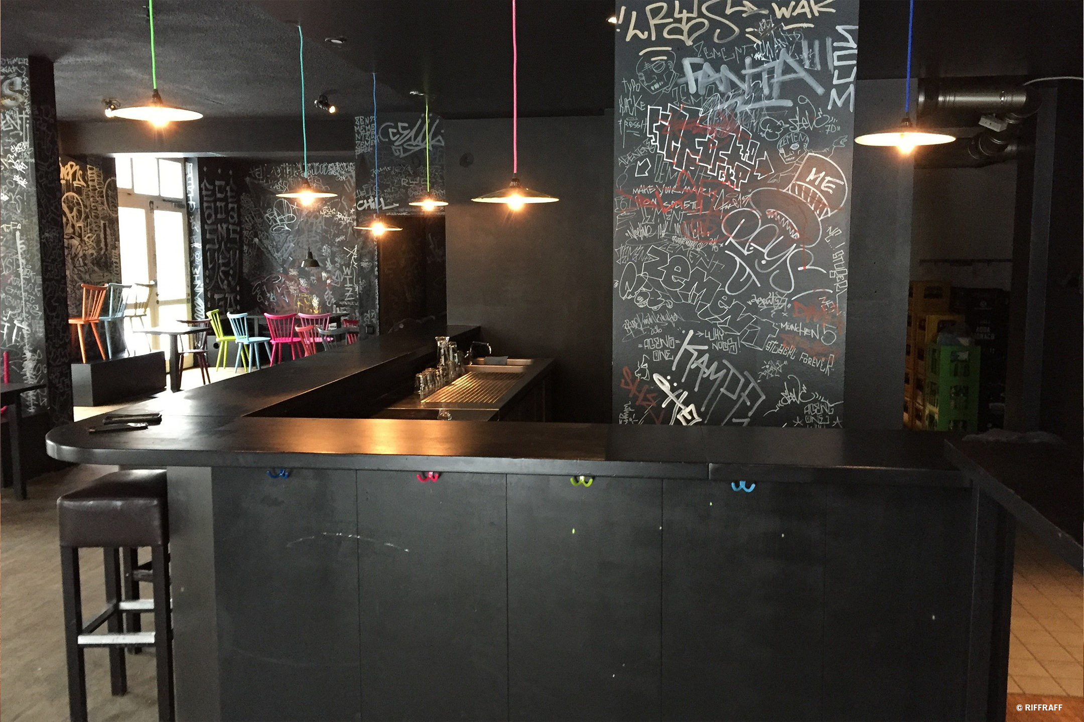 Die Theke einer schwarz gestrichenen Bar mit bunten Leuchten und Stühlen sowie Street Art an den Wänden