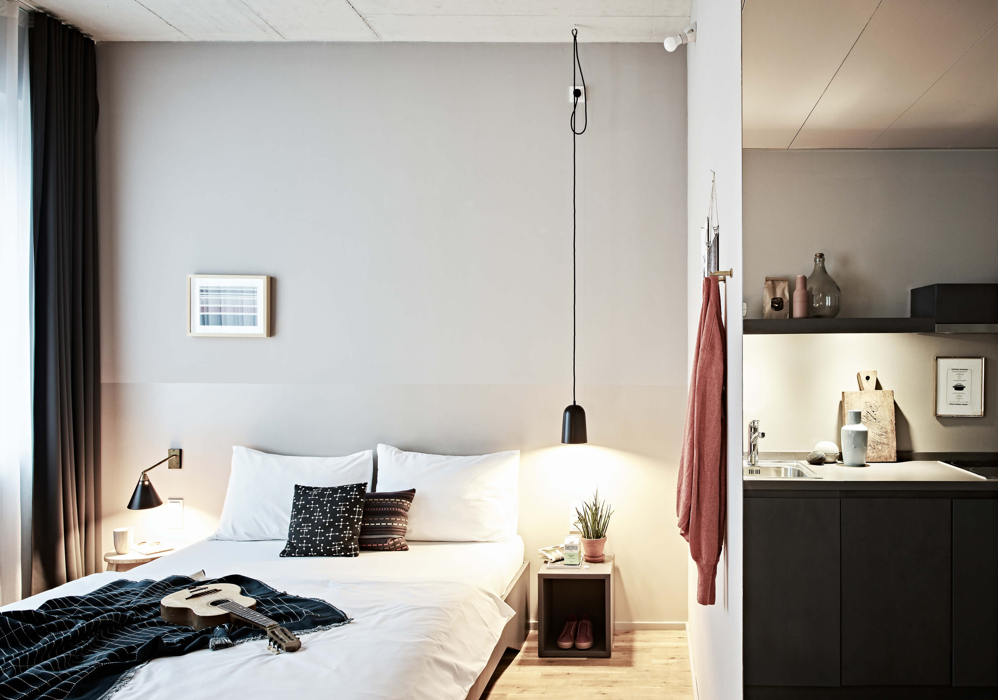 Helles Zimmer mit Holzboden, mittig ein weiß bezogenes Doppelbett und Accessoires, links große Fenster mit Vorhängen, rechts, hinter einer Wand befindet sich eine Kitchenette.