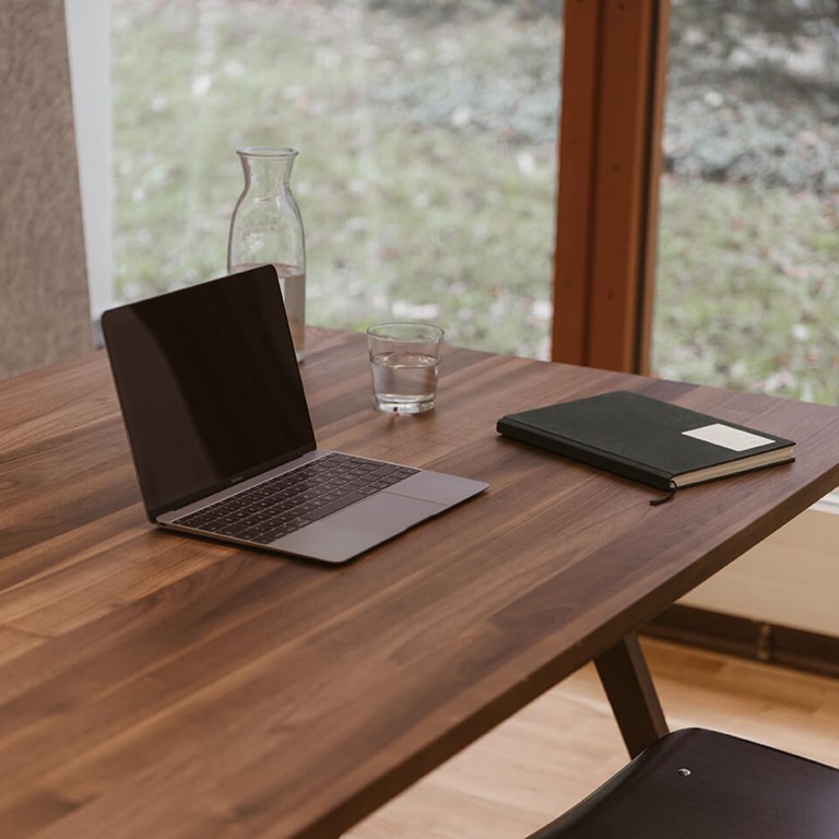 Dunkler Holztische auf hellem Holzboden, davor ein schwarzer Stuhl. Ein Laptop, ein Glas Wasser und ein Notizbuch sind auf dem Tisch. Im Hintergrund ein bodentiefes Fenster mit Blick ins Grüne.