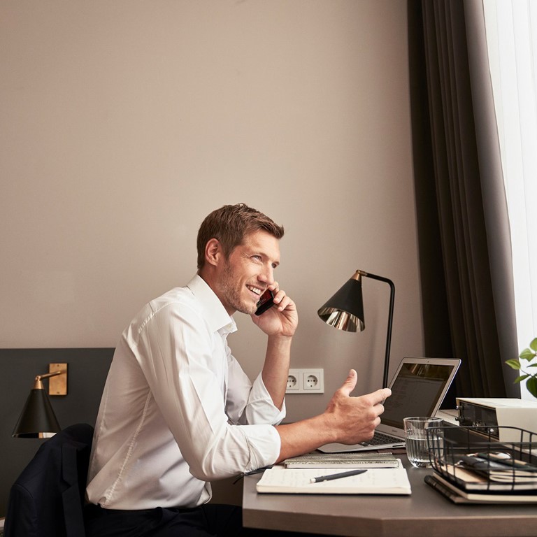 A man sits on the phone at a desk in a hotel room, he smiles and gestures