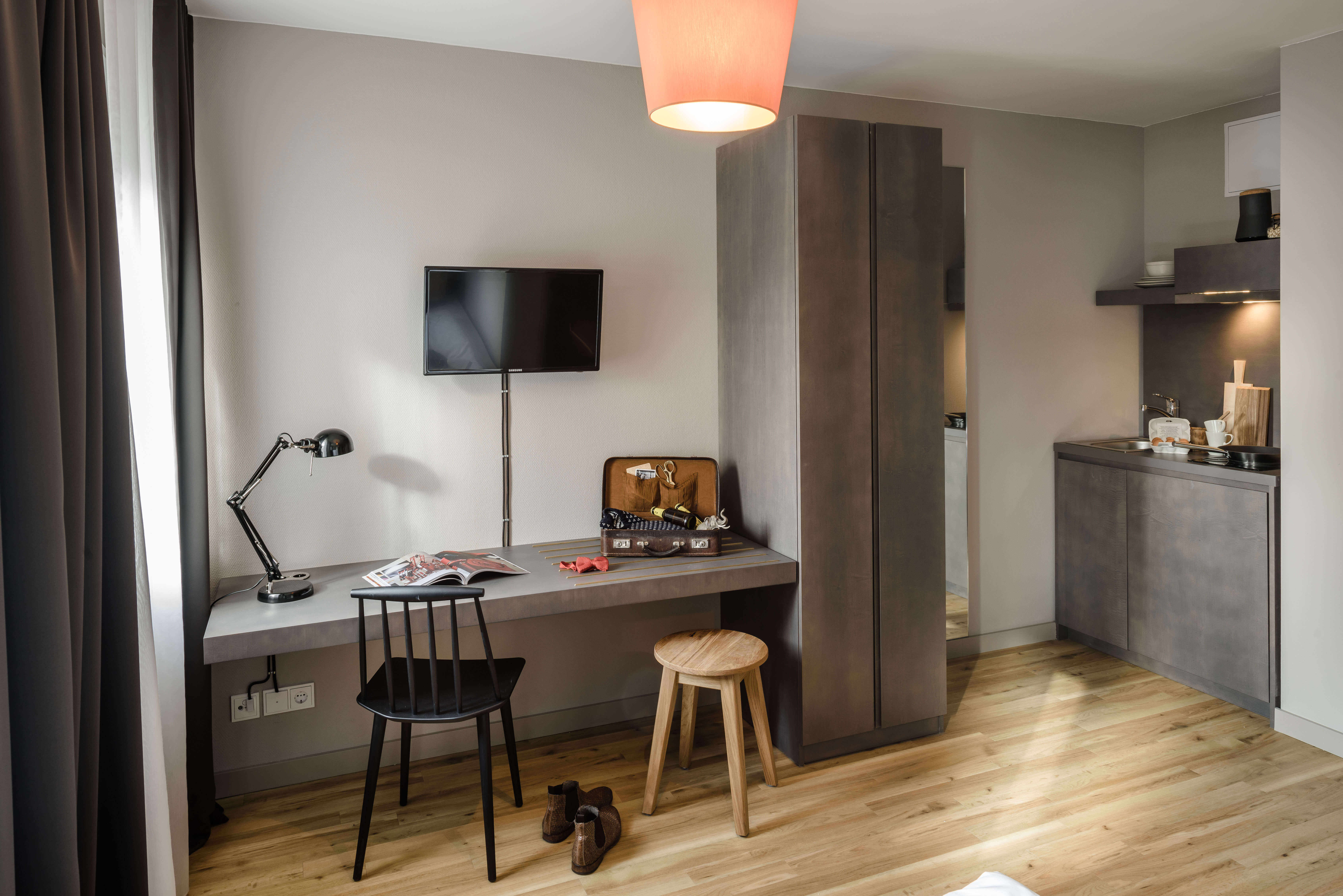 View of a desk with accessories in a room, to the left of it curtains in the cut, in front of it a black chair and a wooden stool. To the right is a kitchenette.