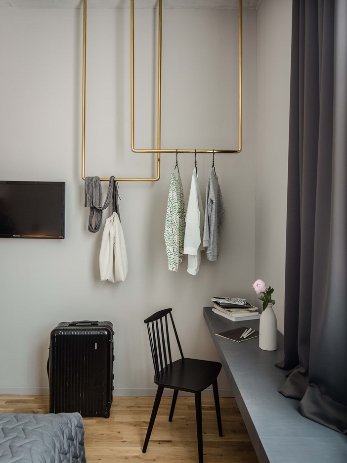 Two gold-coloured, curved bars coming down from the ceiling as a coat rack for various items of clothing. On the left is a flat-screen TV, on the right a long shelf under a large window, in front of which is a black wooden chair.