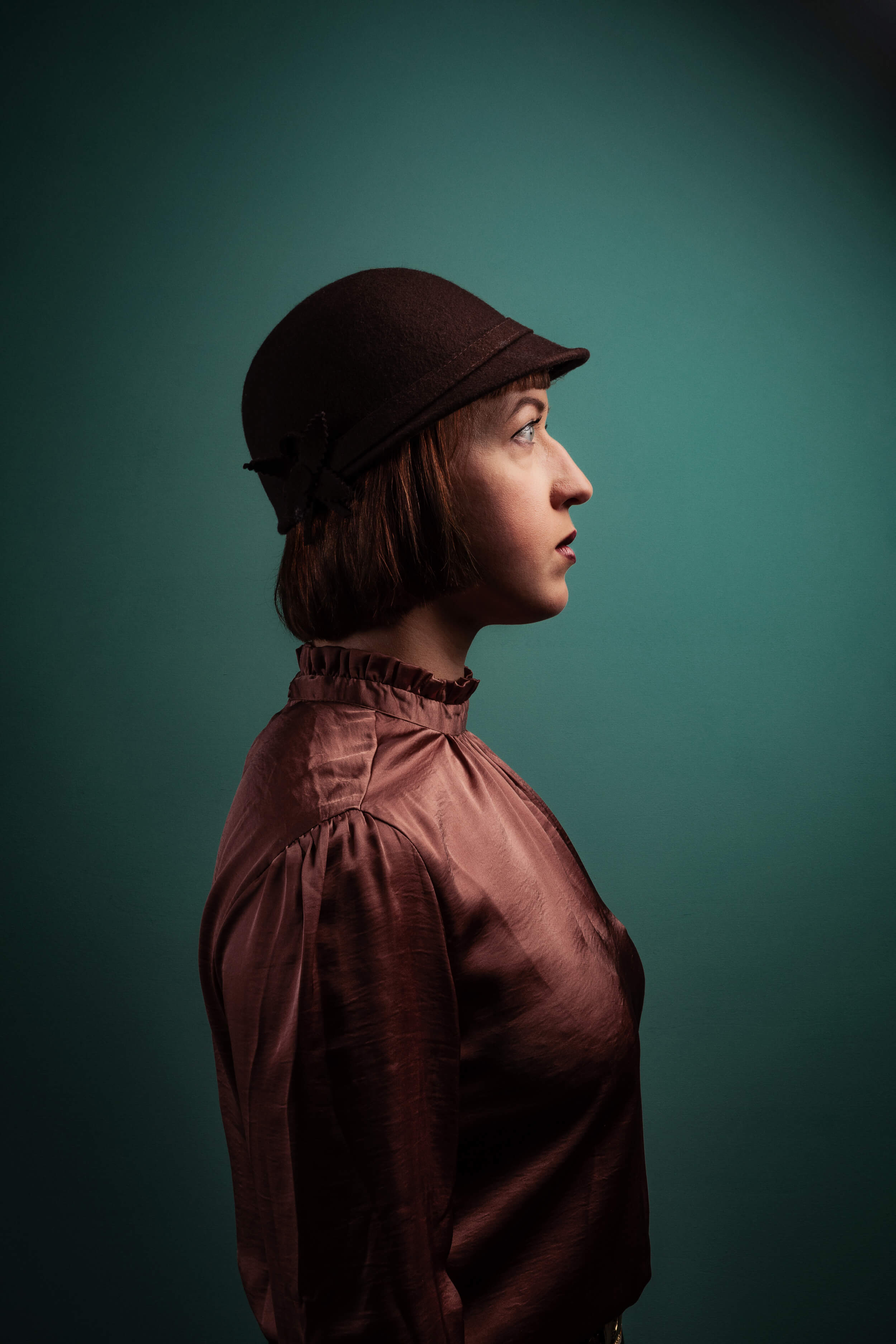 Eine rothaarige Frau mit halblangen Haaren, hochgeschlossener Bluse und Hut steht seitlich vor einem türkisen Hintergrund. Ihr Blick folgt ihrer Standrichtung.