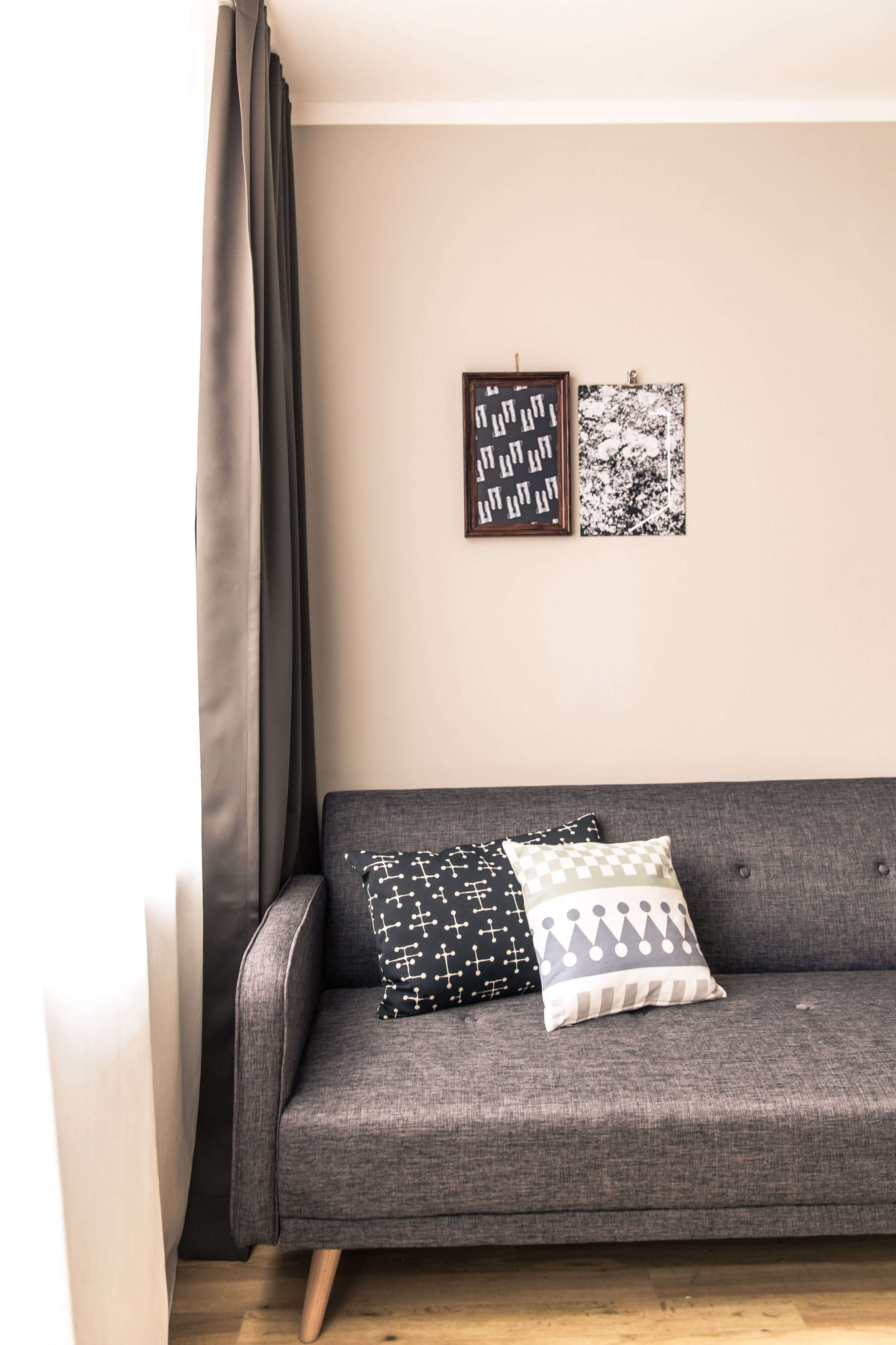 The corner of a dark grey sofa in front of a wall with two pictures, colourful decorative throw cushions on it. A window with long curtains is visible in the cut-out on the right.