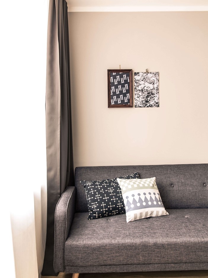 The corner of a dark grey sofa in front of a wall with two pictures, colourful decorative throw cushions on it. A window with long curtains is visible in the cut-out on the right.
