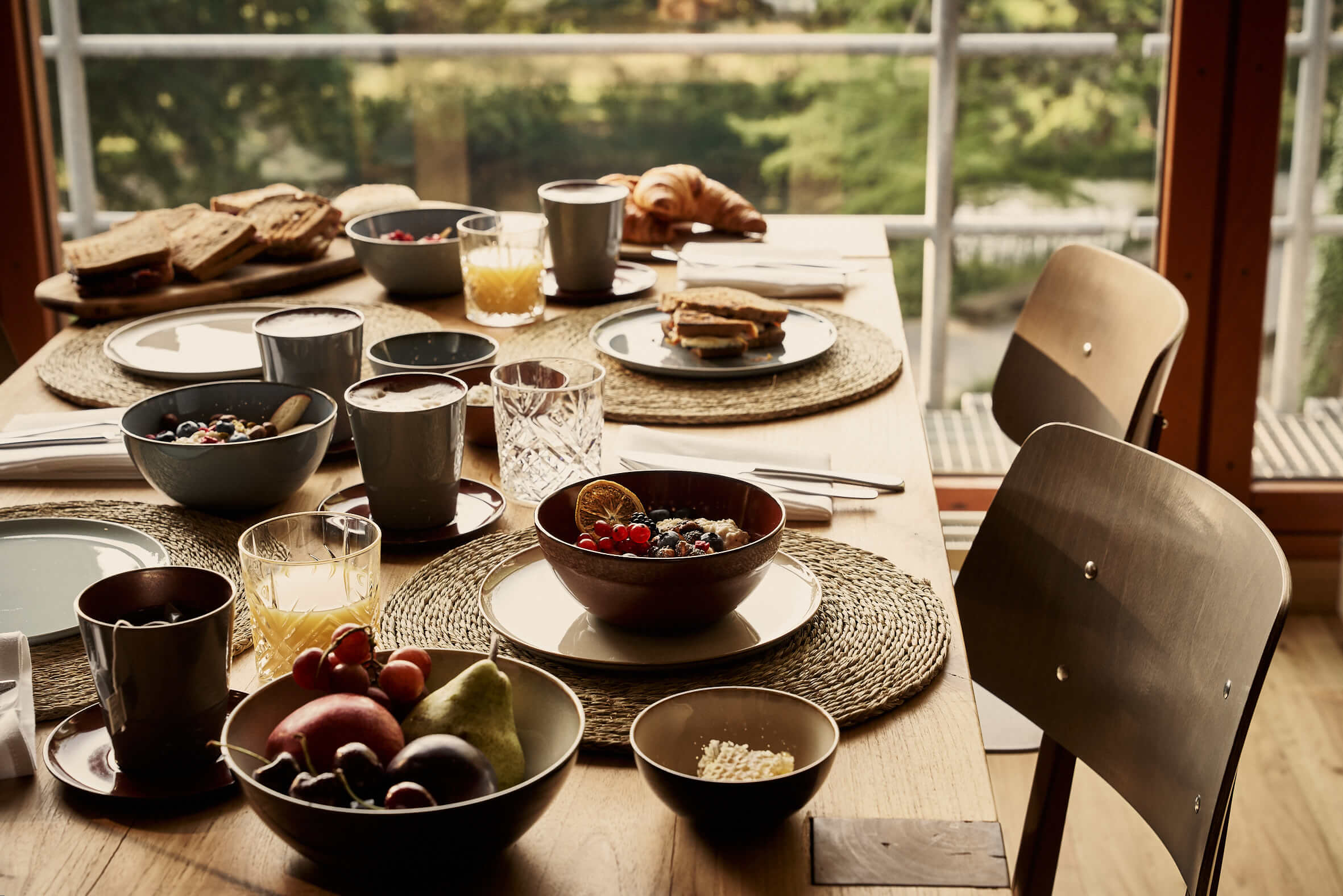 A set wooden table with breakfast ingredients such as porridge, fruit, sandwiches, croissants, and orange juice in front of a large window overlooking a park. 