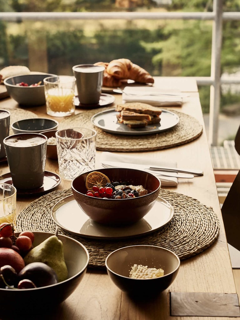 A set wooden table with breakfast ingredients such as porridge, fruit, sandwiches, croissants, and orange juice in front of a large window overlooking a park. 