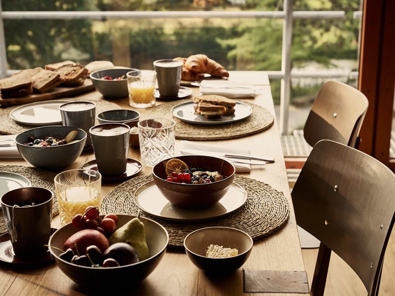 Ein gedeckter Holztisch mit Frühstückszutaten wie Porridge, Früchte, belegte Brote, Croissants, und Orangensaft vor einem großen Fenster, aus dem man einen Park sieht. 