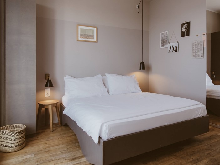 Room with beautiful wooden floor and contrasting walls in grey/old pink, in front a large double bed and various accessories and pictures, on the right a wall mirror.