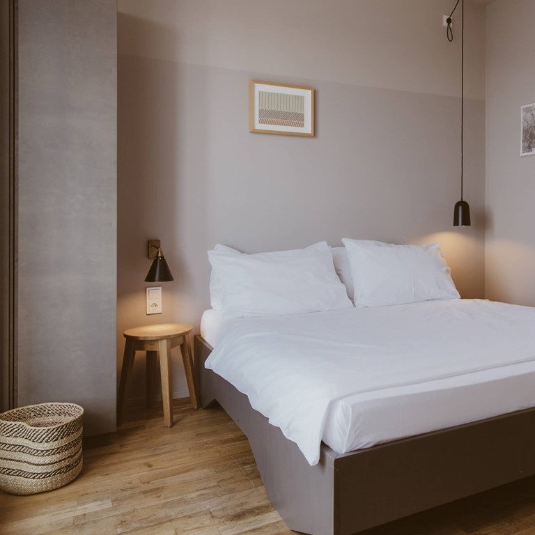 Room with beautiful wooden floor and contrasting walls in grey/old pink, in front a large double bed and various accessories and pictures, on the right a wall mirror.
