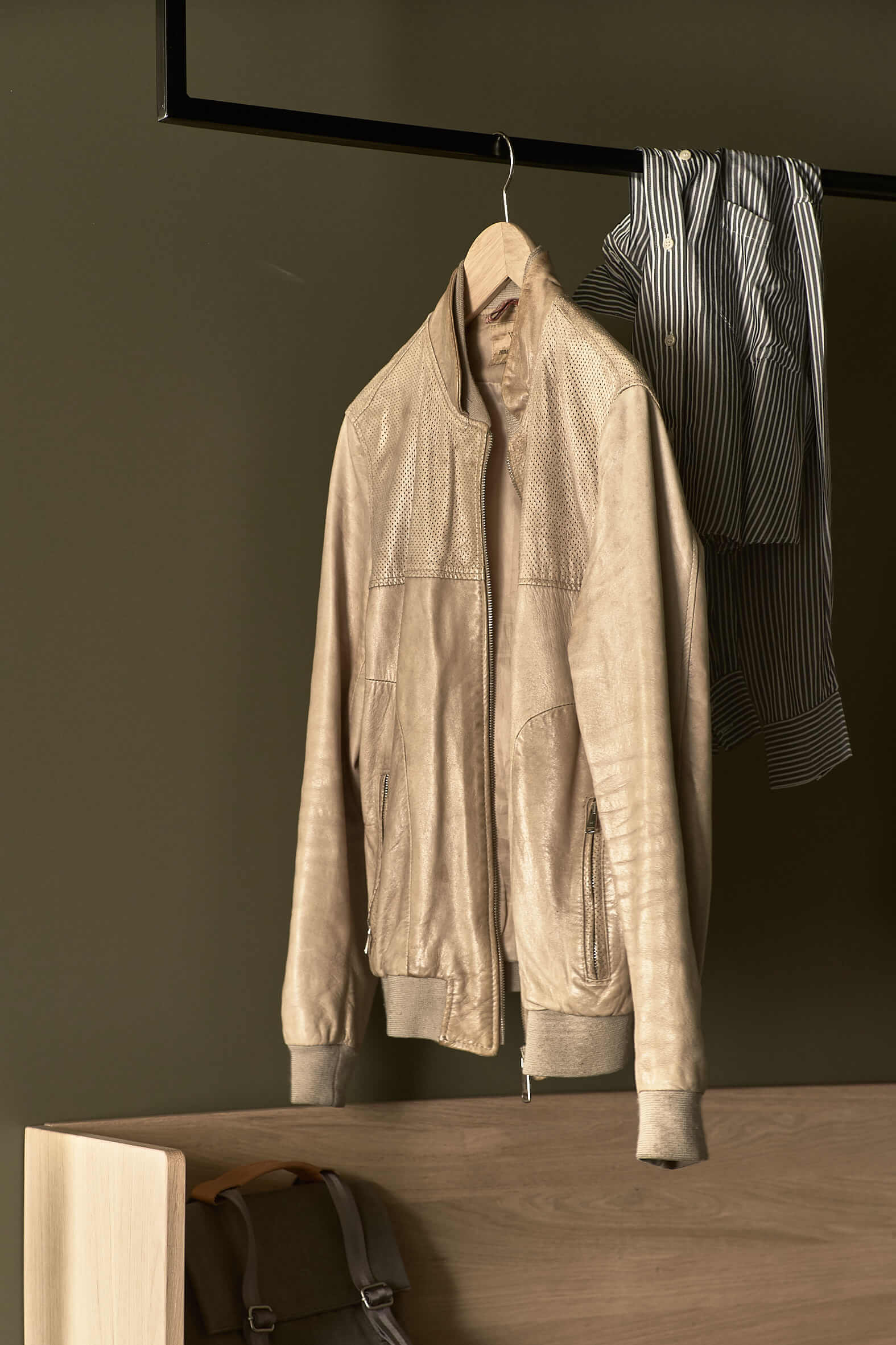 Wooden shelf with a rucksack, above it a solid, angular metal element from which a cream-coloured leather jacket hangs, with a striped shirt lying over the element.