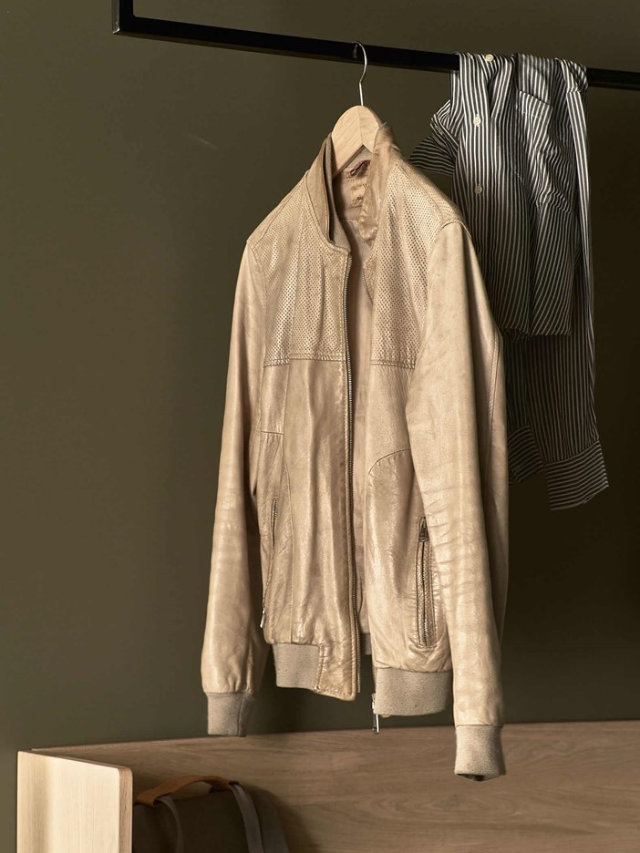 Hölzerne Ablage mit einem Rucksacke, darüber ein massives, eckiges Metallelement an dem eine cremefarbene Lederjacke hängt, über dem Element liegt ein gestreiftes Hemd.