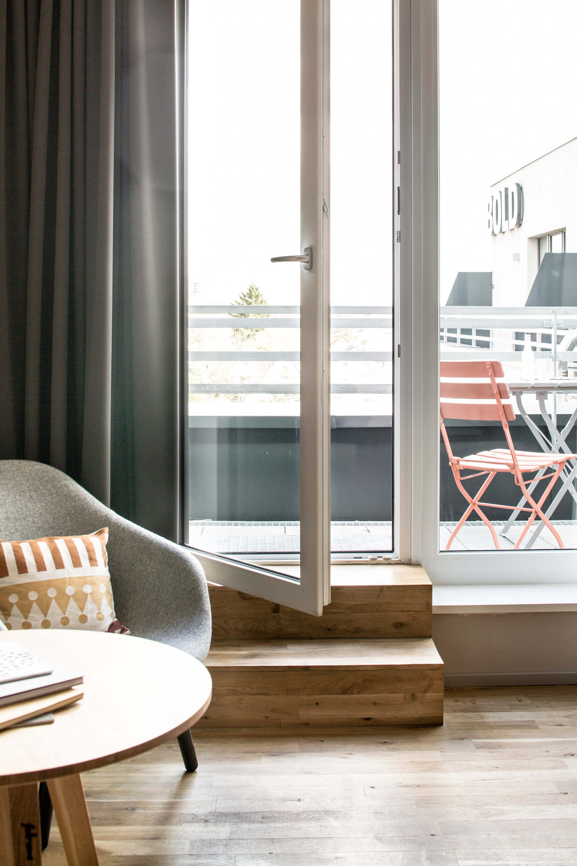 Weiß gerahmte Terrassentür, die über zwei Stufen aus einem Zimmer auf eine Dachterrasse führt. Links ein runder Holztisch mit gemütlichem Sessel, draußen ein bunter Stuhl mit Tisch davor.  