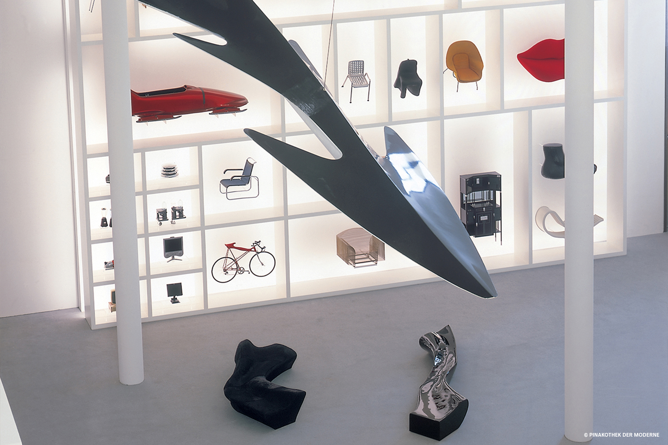 Artworks and product design drafts arranged in a white shelf illuminated from behind, two sculptures in the front, another one suspended from the ceiling