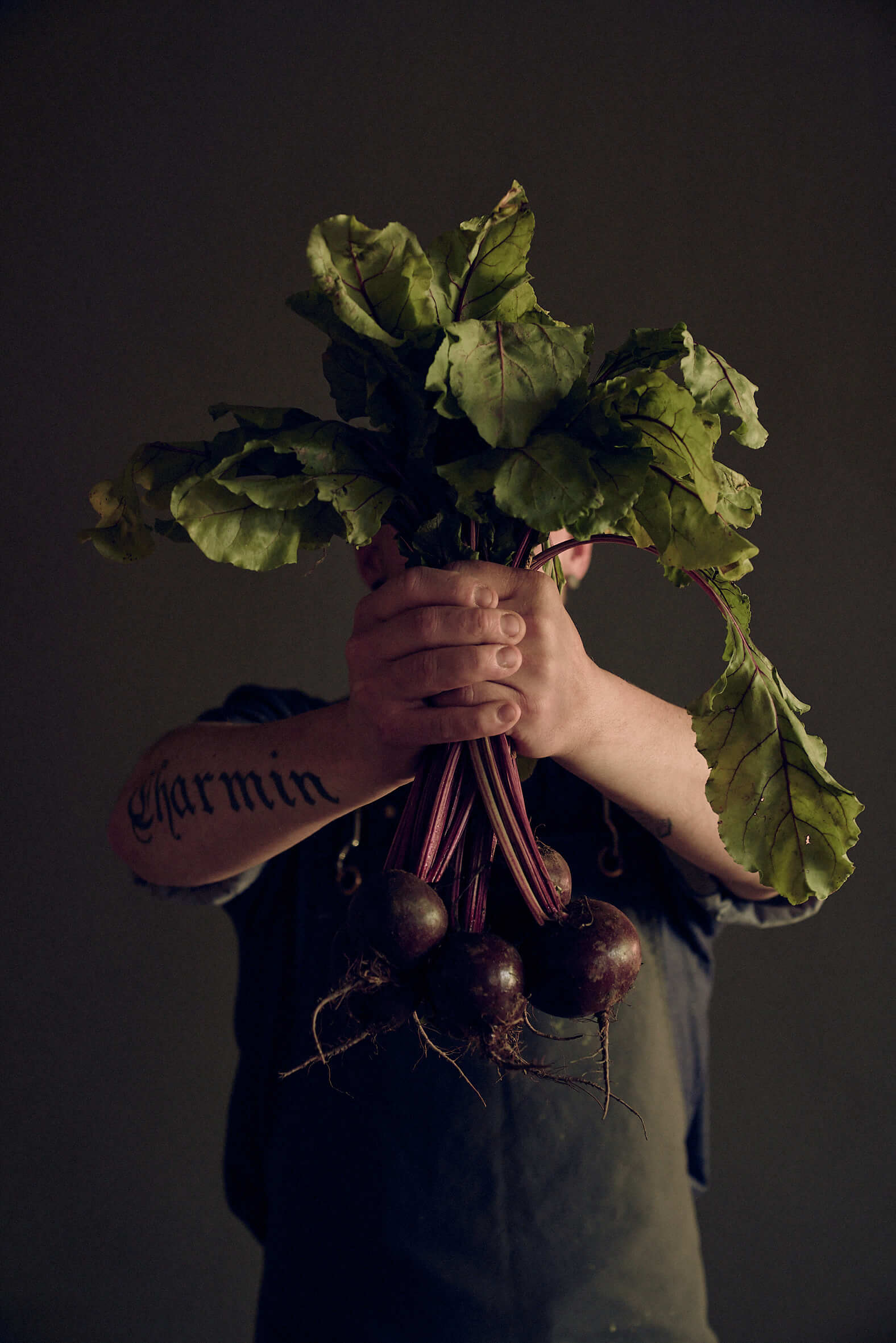 Tätowierter Mann in dunkler Kochschürze hält mit beiden Händen ein Bündel rote Beete in die Kamera. 