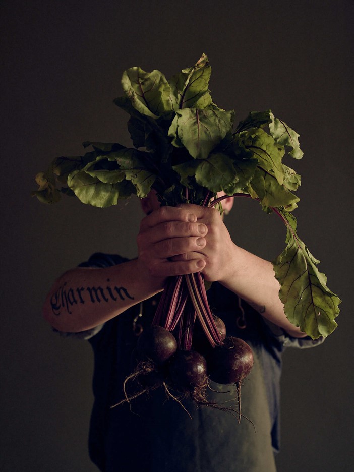 Tattooed man in dark cooking apron holds a bunch of beetroot with both hands into the camera. 