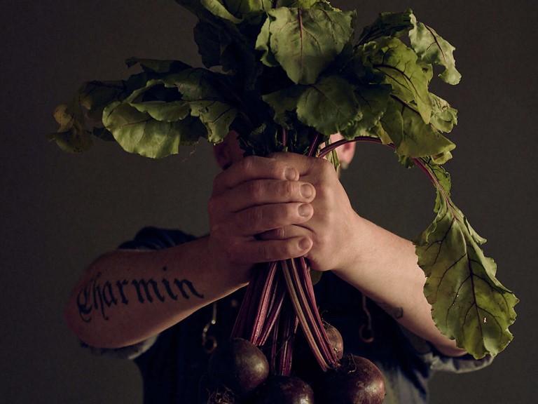 Tattooed man in dark cooking apron holds a bunch of beetroot with both hands into the camera. 