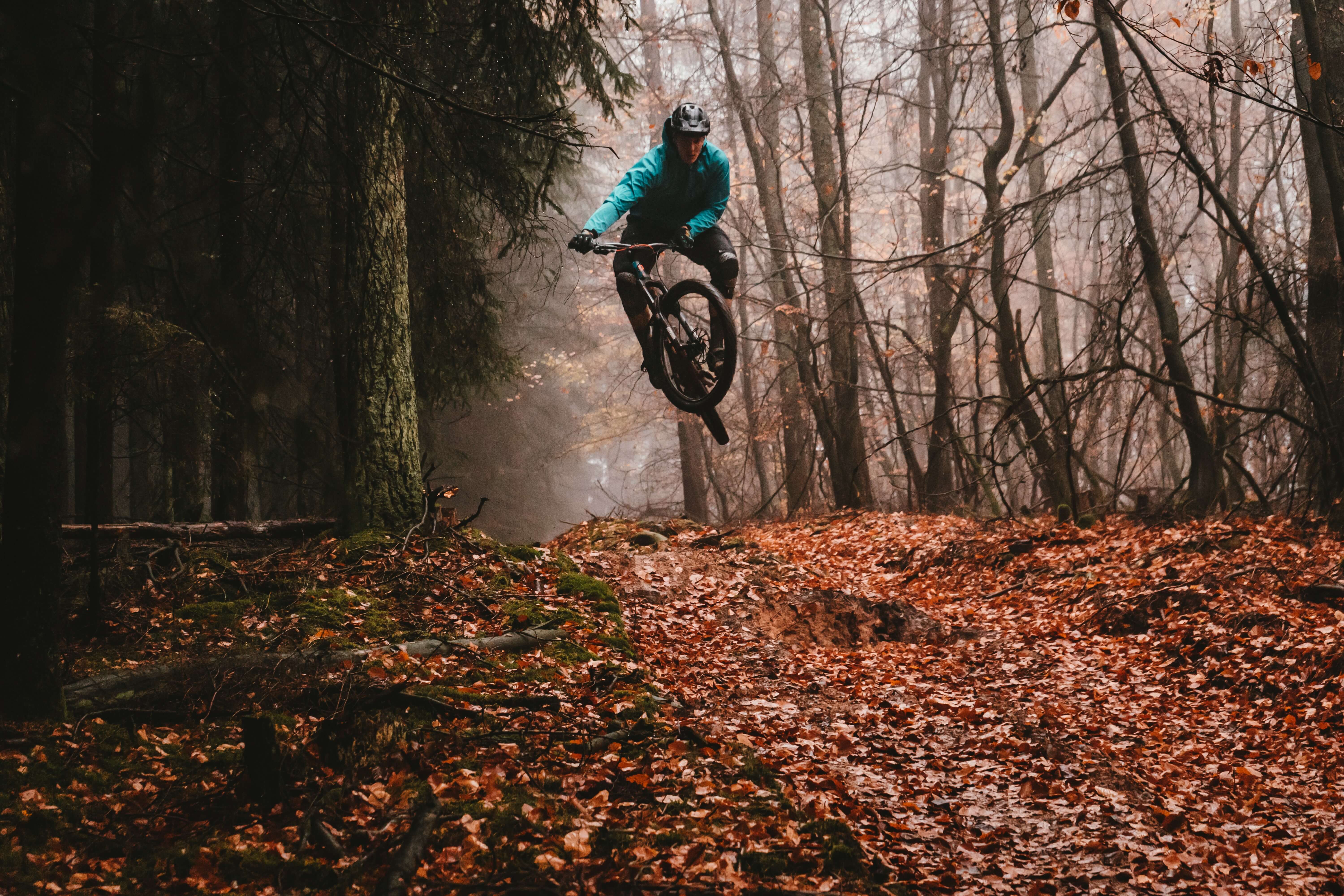 Ein Mann in Outdoor Klamotten und Helm springt mit einem Mountainbike durch eine herbstliche Waldlandschaft. Im Vordergrund sind braune Blätter auf dem Boden zu sehen. Zwischen den Bäumen ist Nebel.