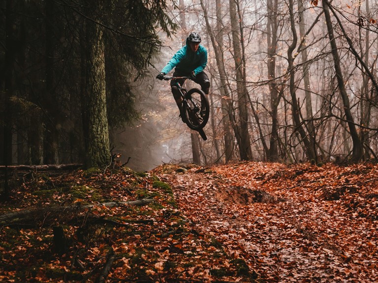A man in outdoor clothes and helmet jumps on a mountain bike through an autumnal forest landscape. Brown leaves can be seen on the ground in the foreground. There is fog between the trees.