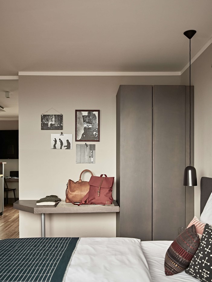 Ein gemütliches Doppelbett mit Accessoires und einem grauen Kleiderschrank dahinter sowie eine Ablage mit Taschen. An der Wand hängen gerahmte, schwarz-weiße Bilder.