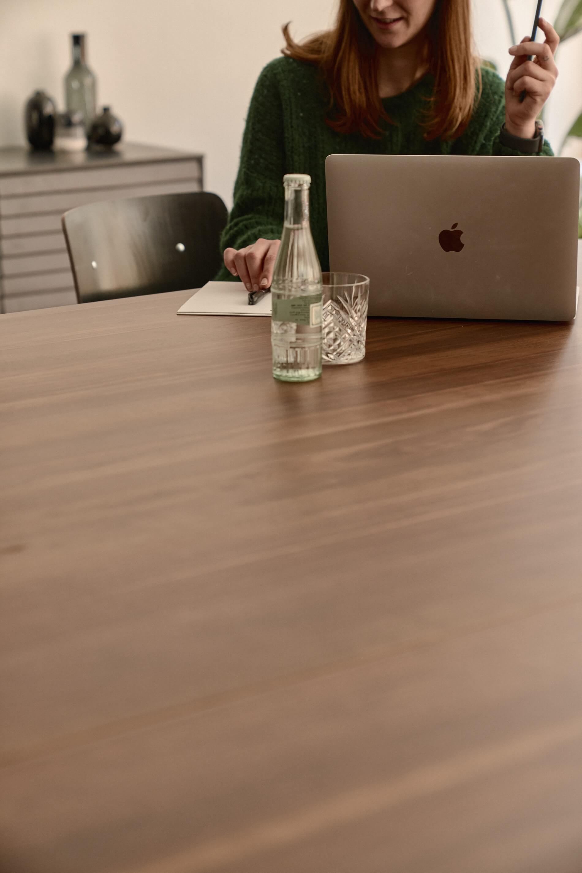 A woman is sitting at a table in front of a laptop. She has a pen in her hand and a bottle of water and a glass in front of her.
