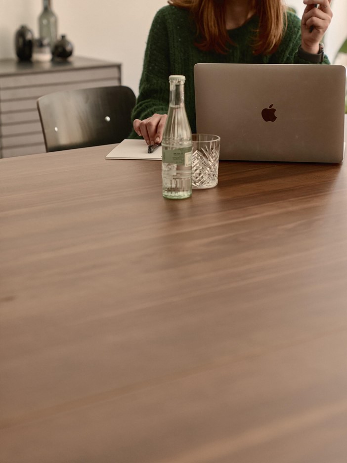 Eine Frau sitzt an einem Tisch vor einem Laptop. In der Hand hält sie einen Stift und vor ihr steht eine Flasche Wasser sowie ein Glas. 
