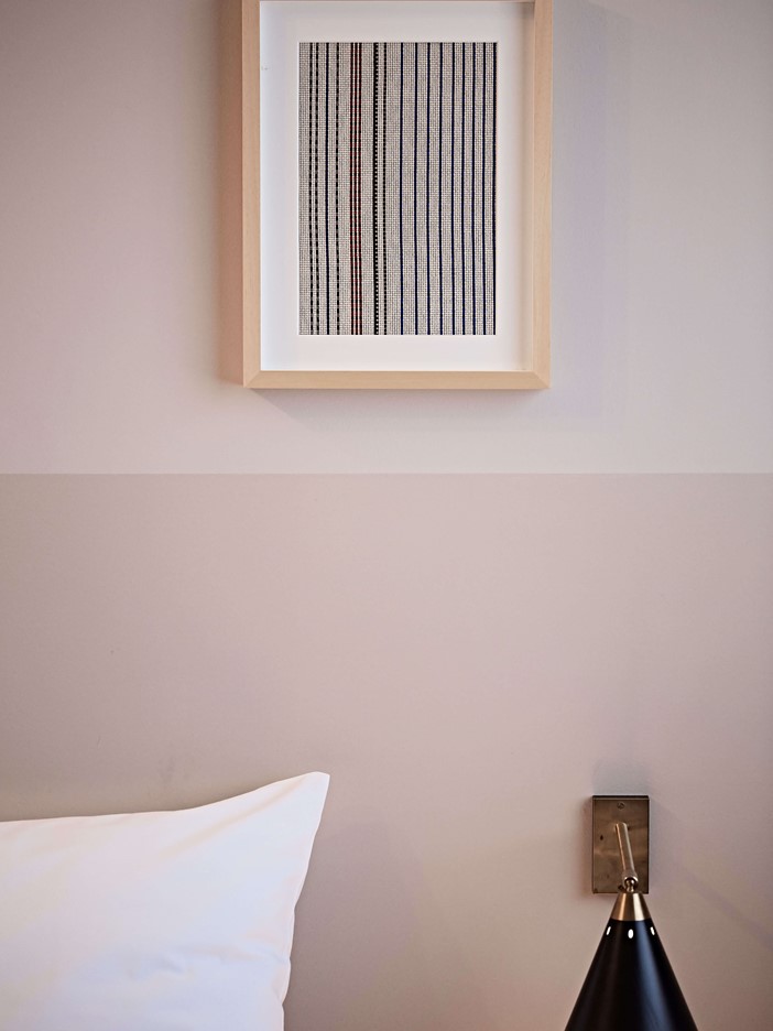 Ein minimalistisches Bild mittig an einer zweifarbigen Wand, darunter rechts eine schwarze Leseleuchte, im Anschnitt links ein weißes Kissen.