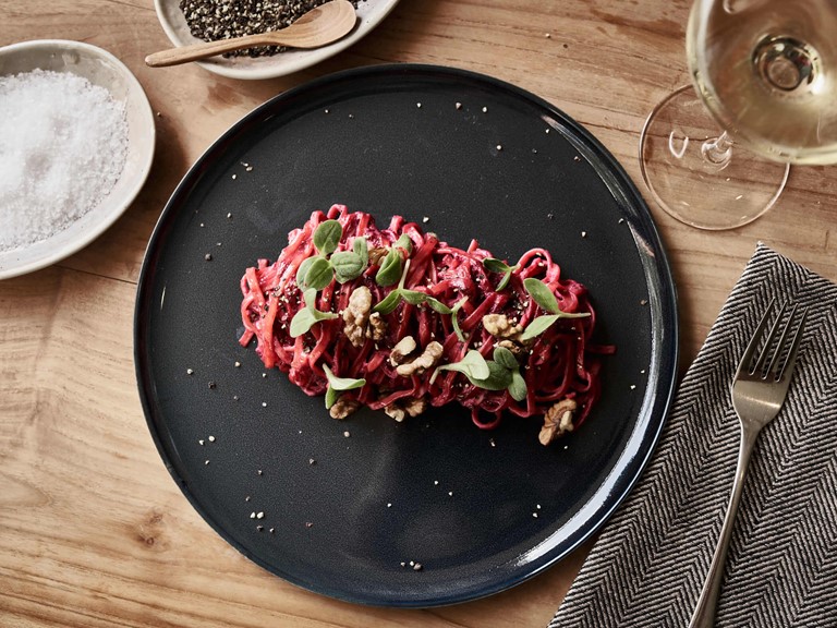 Bordeaux-coloured ribbon noodles garnished with green leaves and nuts, beautifully arranged on a dark plate. Next to it a fork on a cloth napkin and a glass of white wine.