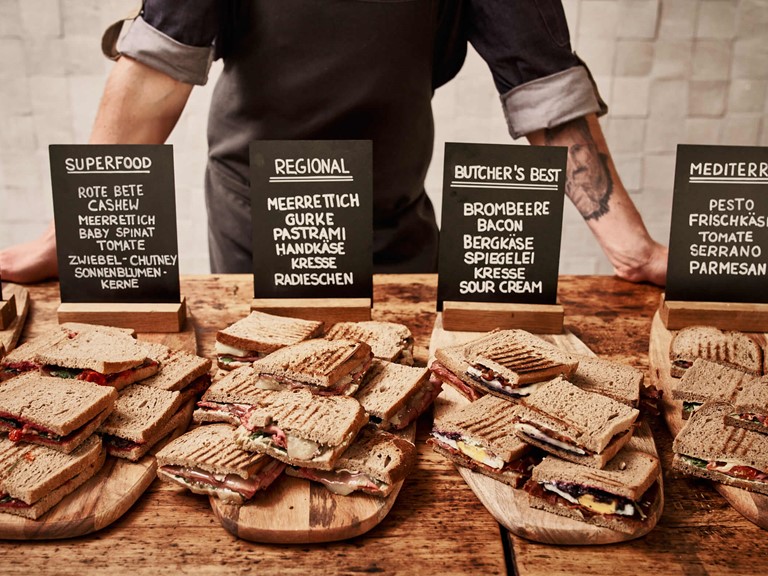 Große Auswahl verschieden belegter Sandwiches, die auf Holzbrettern liegen, dahinter beschriftete Tafeln mit Zutaten und die Arme eines Mannes, der sich aufstützt.