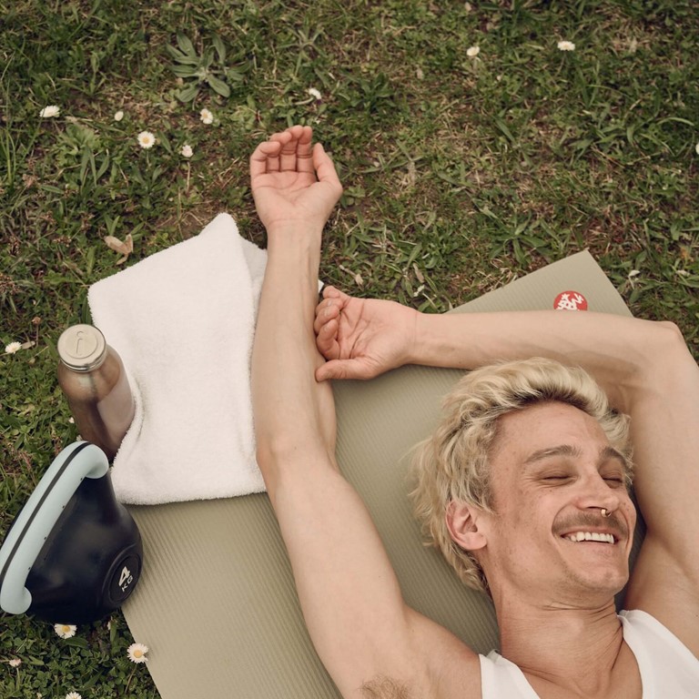 Junger Mann mit Bart und Piercing liegt im Park des Bold Campus auf einer Yogamatte und lächelt