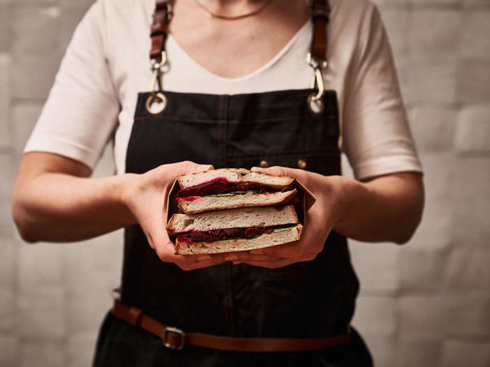 Person in a chef's apron and white T-shirt holding a sliced, thick sandwich with both hands towards the camera.