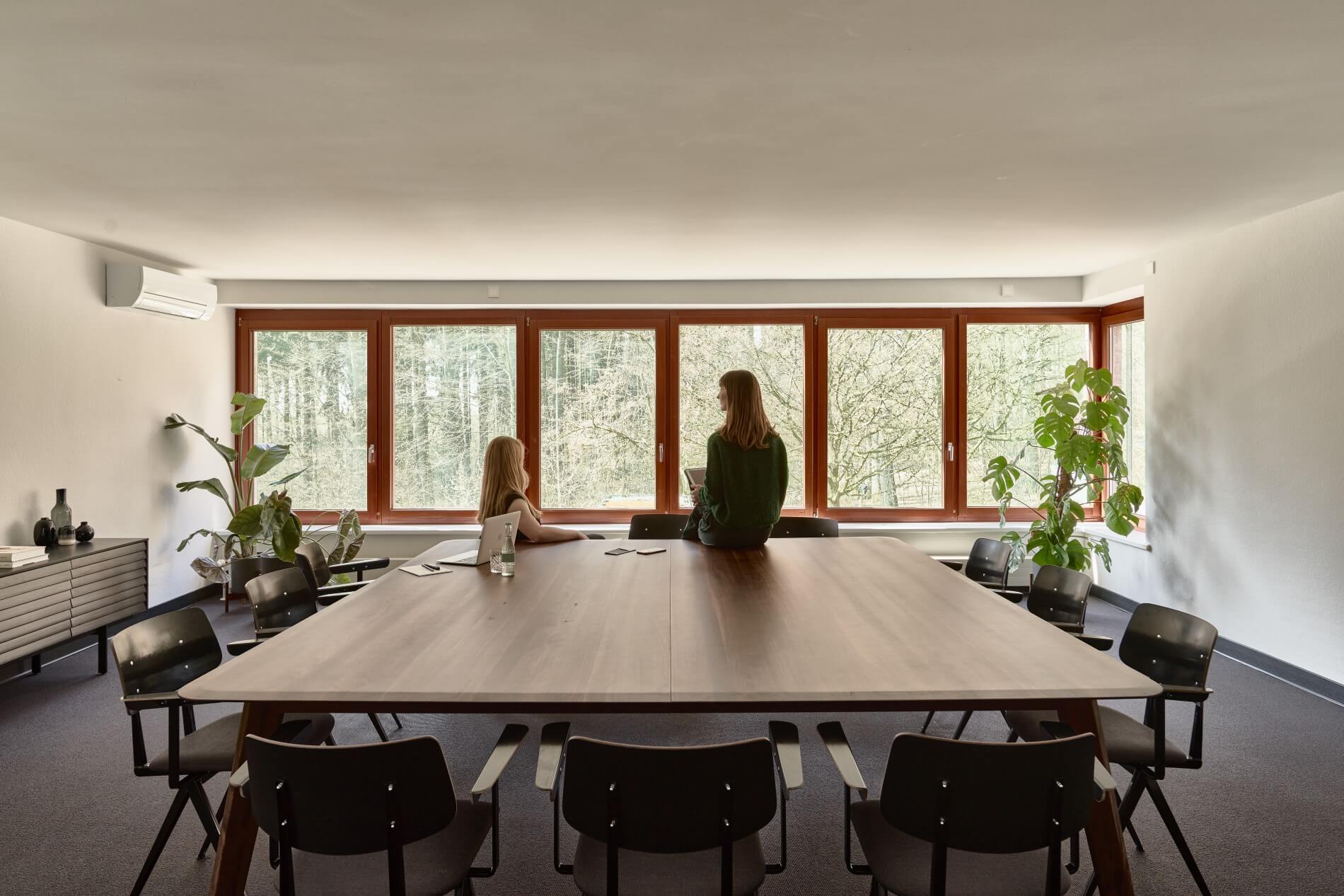 A woman sits on a table in one of the meeting rooms at the Bold Campus and looks out into the forest. Another woman sits on a chair next to her and also looks out. On the table is a laptop, notes and water. Chairs are set up around the table.