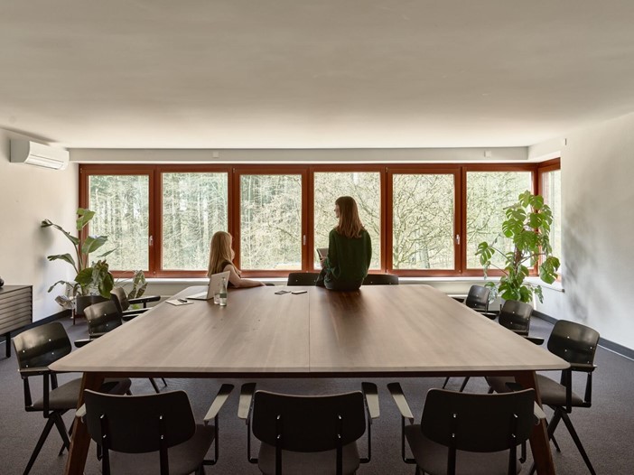A woman sits on a table in one of the meeting rooms at the Bold Campus and looks out into the forest. Another woman sits on a chair next to her and also looks out. On the table is a laptop, notes and water. Chairs are set up around the table.