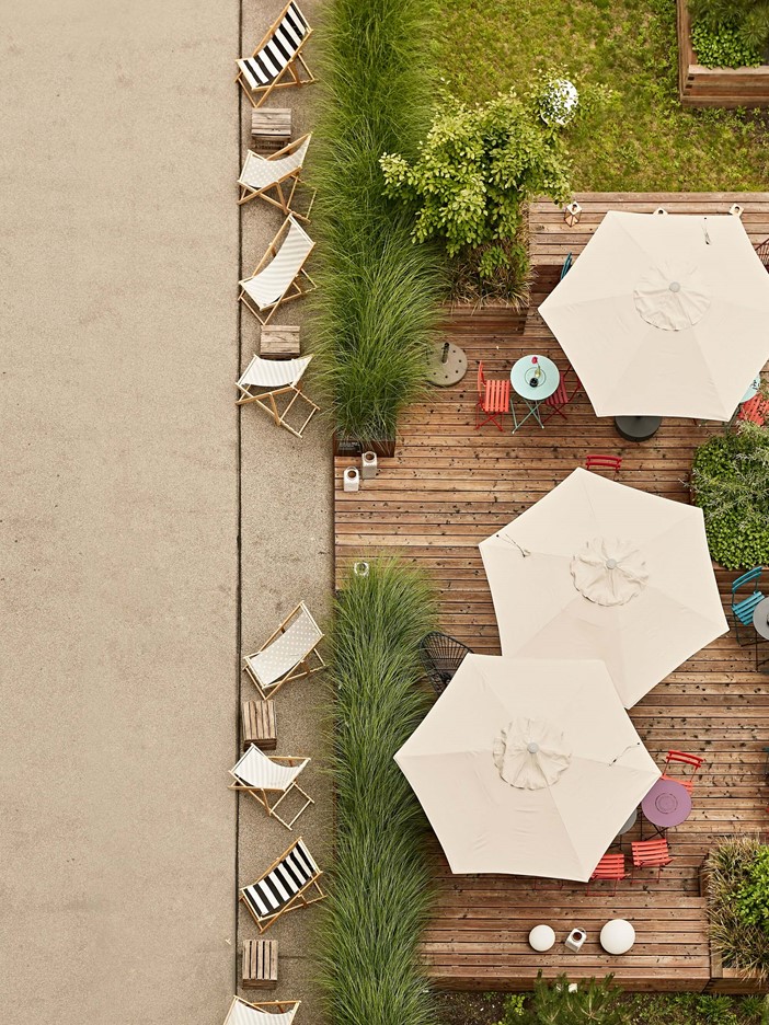 View from above onto a terrace with large white parasols, various colourful chairs and deckchairs and many green plants. 